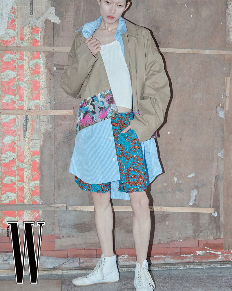 An adult female model in a tan jacket, white top, and patchwork skirt poses indoors against a distressed wall for a fashion editorial.