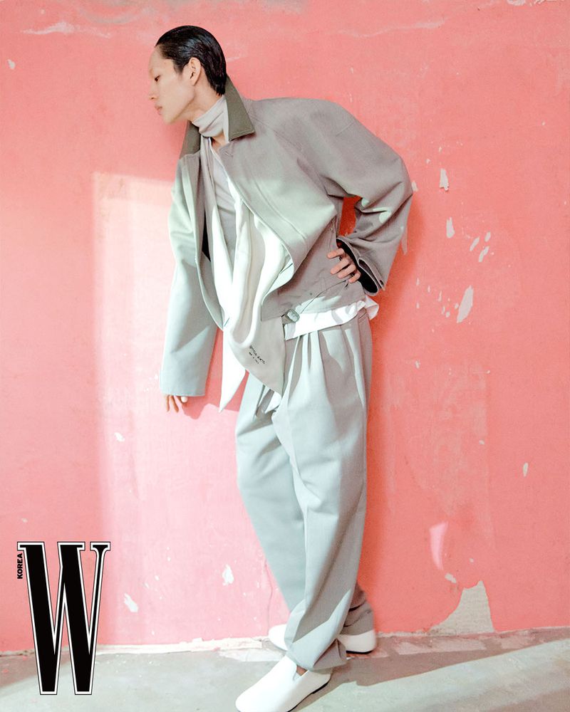 Model wearing grey layered jacket and trousers leaning against a textured pink wall for W Korea editorial.