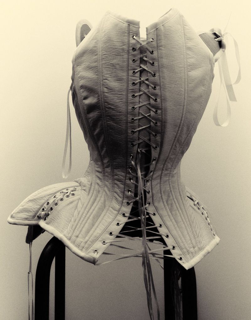 A structured off-white leather corset with lace-up back detailing and boning, displayed on a dark stand against a pale backdrop.