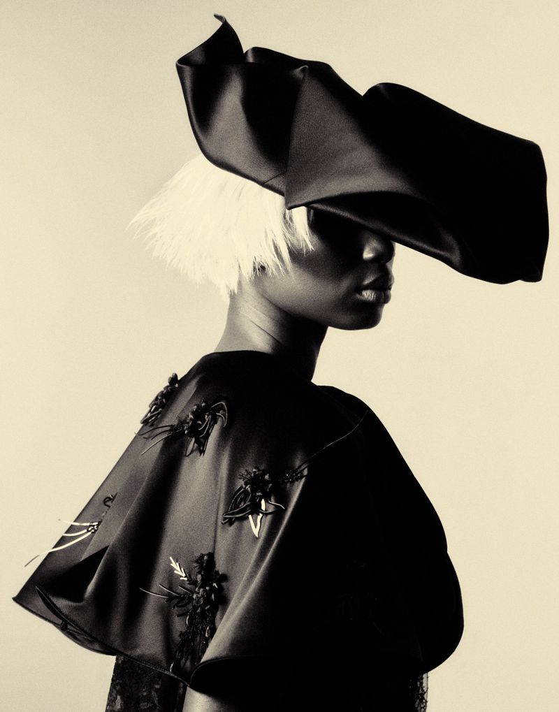 Model Mameanta Wade in a sculptural black headpiece, feathered white hair, and an embellished satin cape against a studio backdrop.