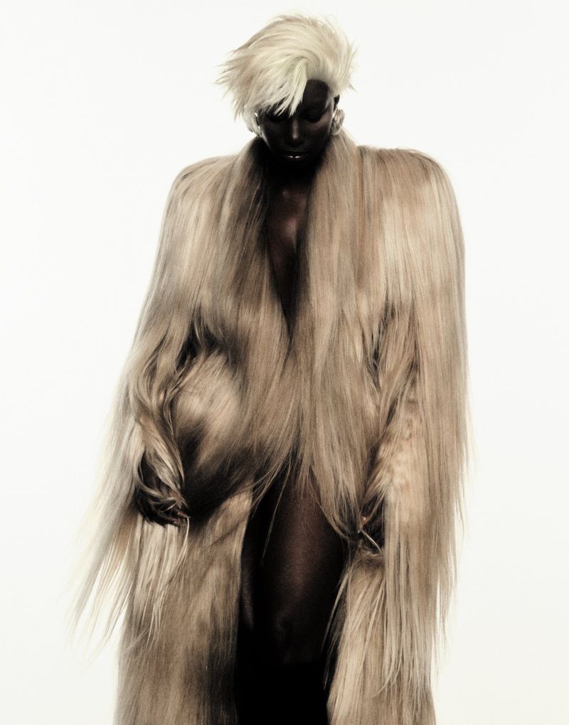 Studio shot of a Black adult female model with dyed white hair wearing a long furry tan coat against a white backdrop for a fashion editorial.