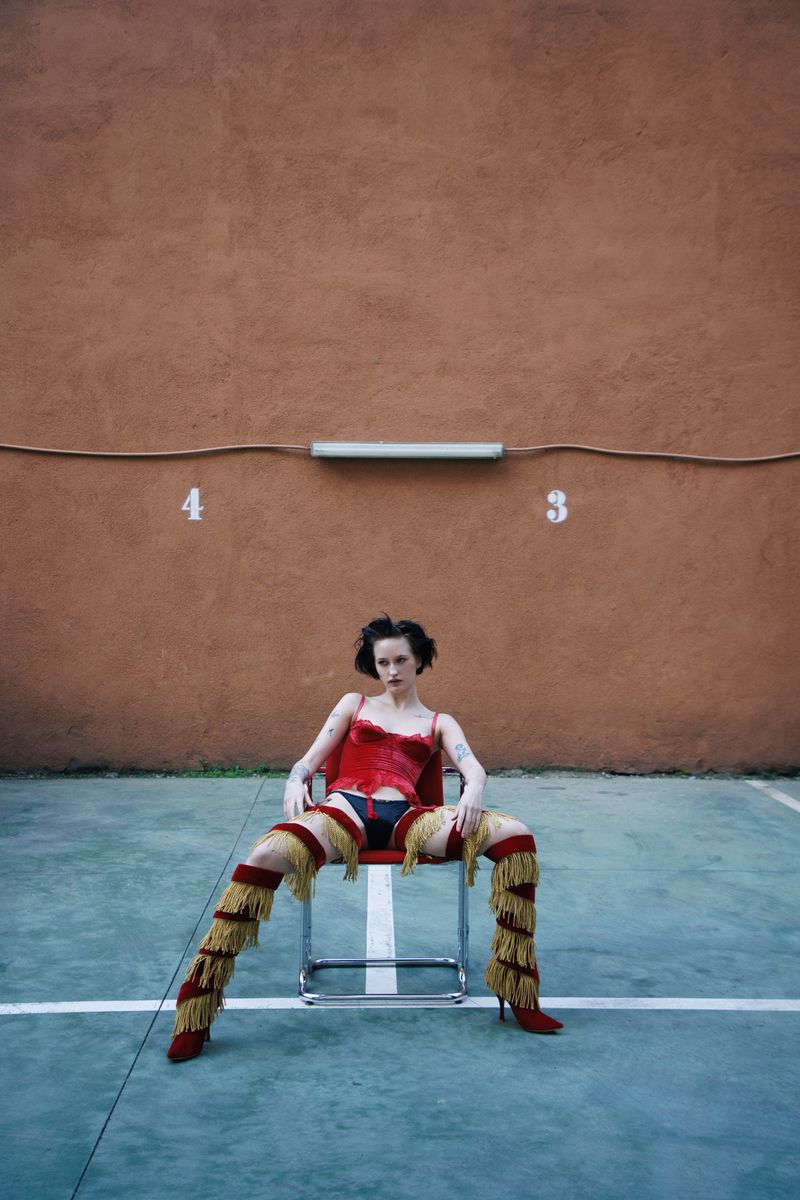 Woman in a red corset and fringe thigh-high boots sitting wide-legged on a chair outdoors in front of a textured terracotta wall.