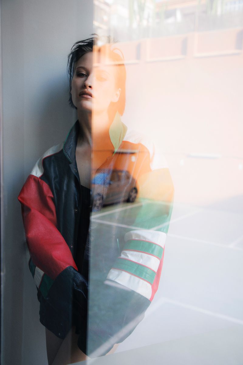 Model with slicked hair wearing an oversized colorblock leather jacket viewed through a reflective glass window.