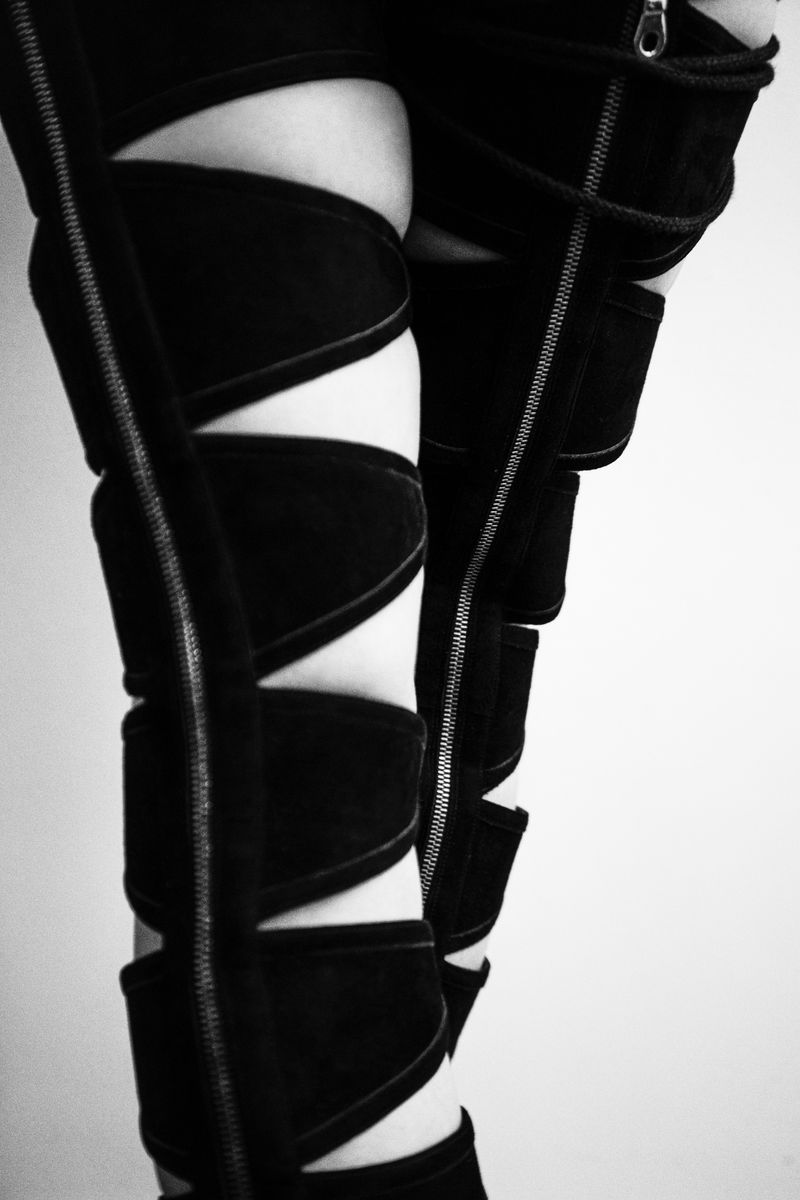 Close-up of legs wearing black cutout strapped boots with vertical metal zippers against a plain light studio backdrop.