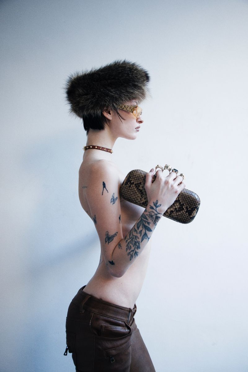 Side profile of a tattooed model wearing a large fur hat, yellow-tinted sunglasses, brown leather pants, and holding a snakeskin clutch.