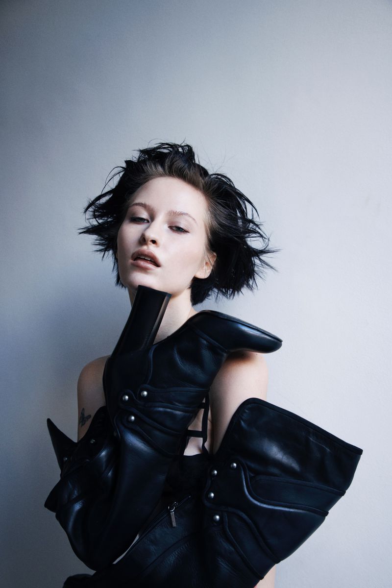 Woman with short tousled dark hair resting her chin on a black studded leather high-heeled boot against a grey background.