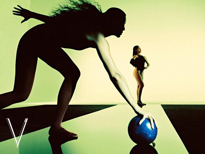 Silhouette of a woman reaching towards a blue ball in a stylized studio with another model standing in the background.