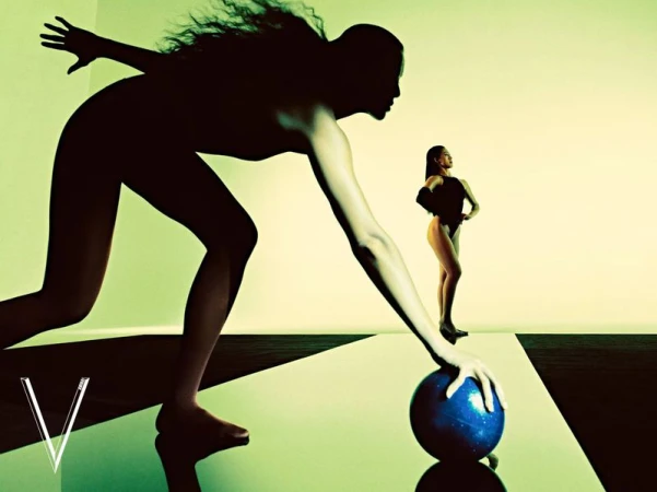 Silhouette of a woman reaching towards a blue ball in a stylized studio with another model standing in the background.