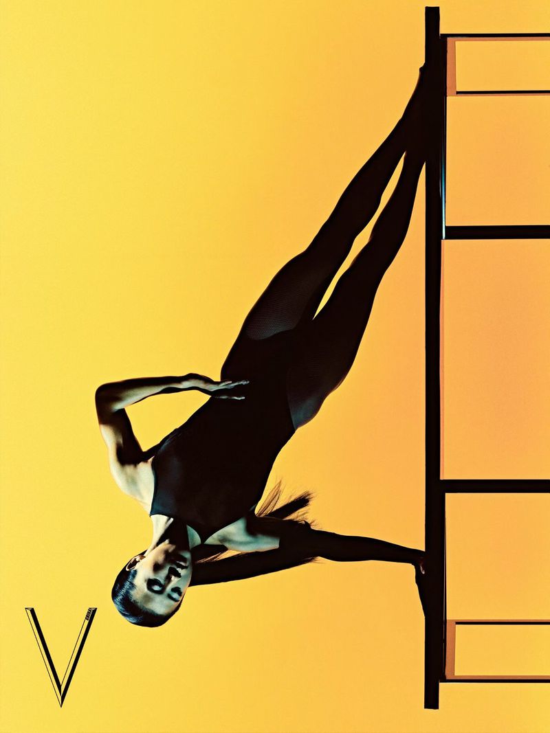 Woman in a black bodysuit posing inverted against a geometric frame on a vibrant yellow gradient background.