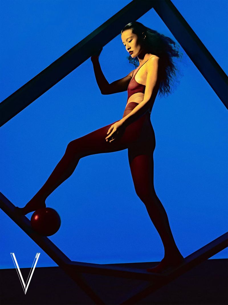 Model in a burgundy bodysuit posing dynamically within a dark geometric frame against a vibrant blue studio backdrop.