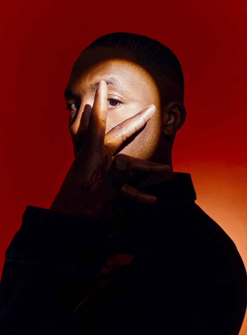 A close-up portrait of a Black man holding his hand in front of his face under a focused beam of light against a deep red background.