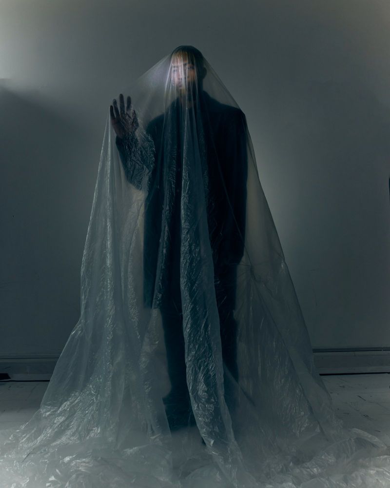 James Massiah standing draped entirely beneath a large translucent plastic sheet with one hand raised in a dim studio.