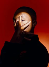 A close-up portrait of a Black man holding his hand in front of his face under a focused beam of light against a deep red background.
