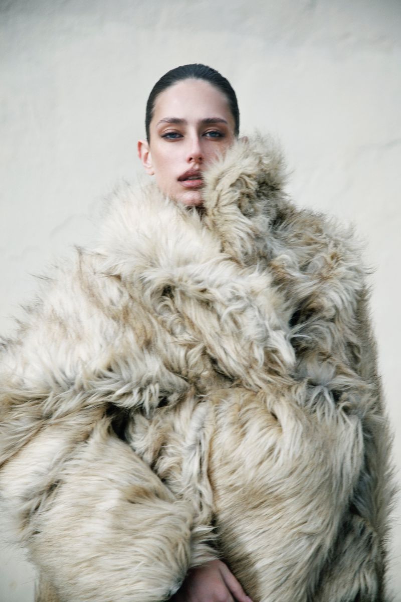 Adult woman wearing a large, beige fur coat against a neutral background. She has wet hair and a neutral expression.