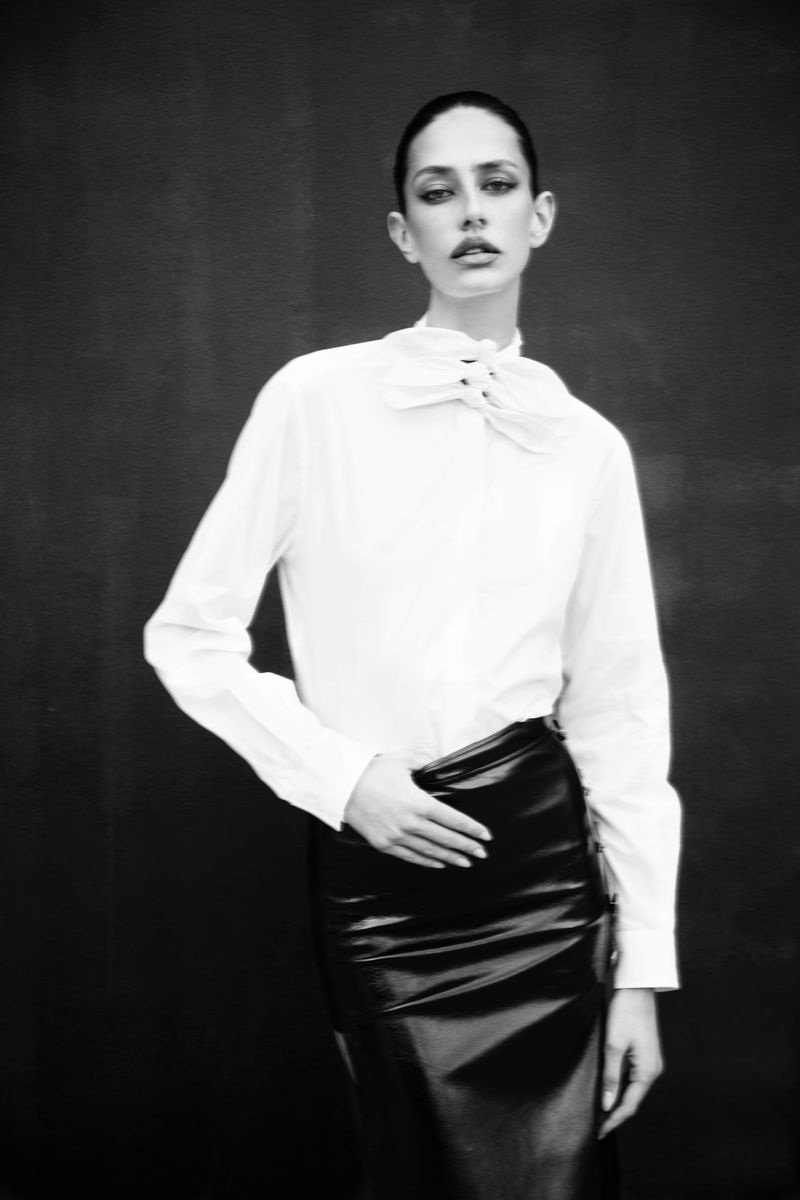 Model Nora Vara poses in a white high-neck bow blouse and black high-waisted skirt against a dark seamless backdrop.
