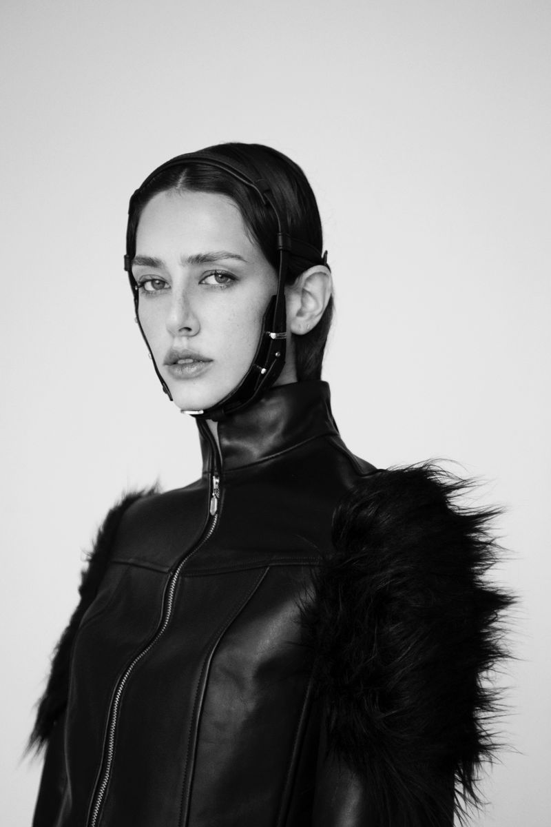 Black and white portrait of model Nora Vara in a high-collar leather jacket with fur shoulders and a strapped headpiece.