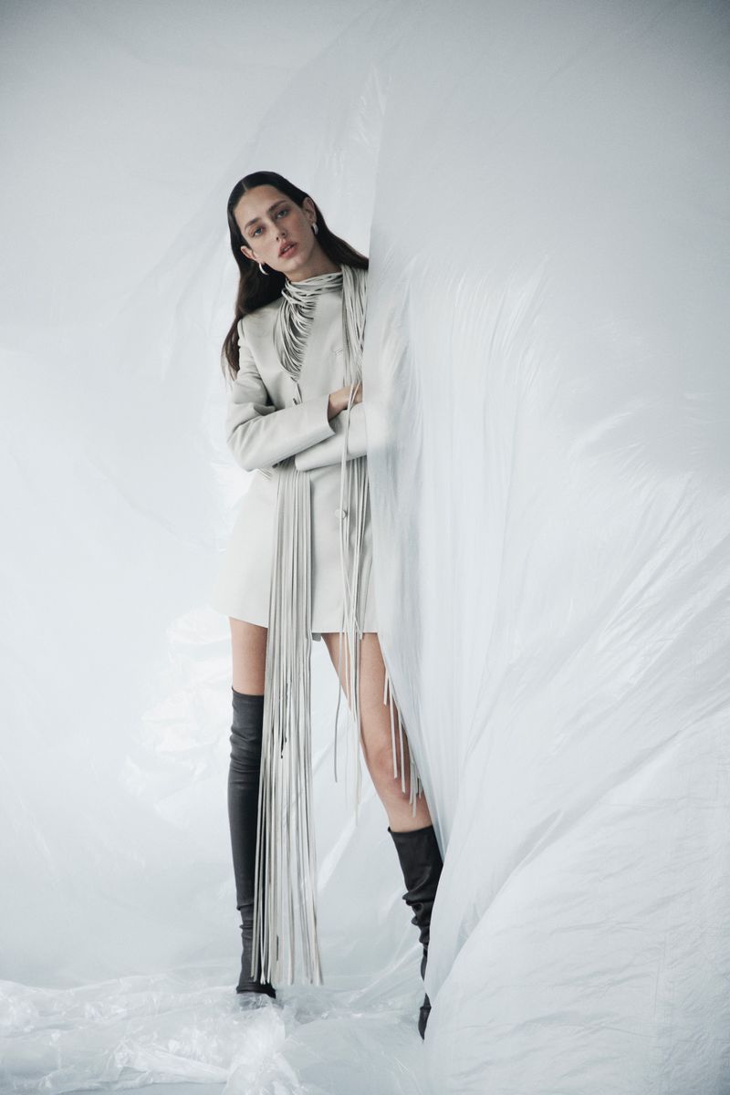 Model Nora Vara wearing an off-white fringe jacket and black thigh-high boots posed behind translucent white plastic sheeting.