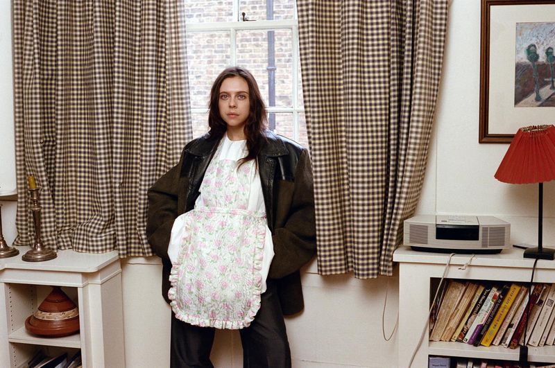 A woman poses in front of a window with checkered curtains. She is wearing an apron and a large jacket in an indoor setting.