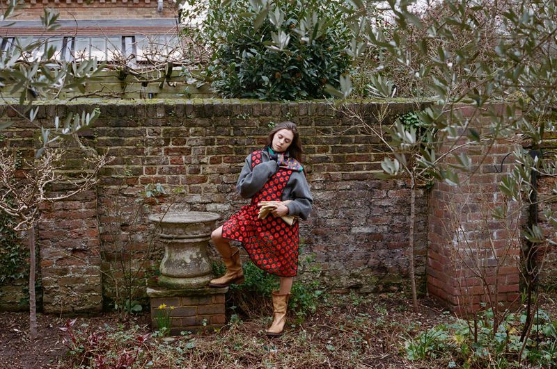Bel Powley in a red patterned dress, grey sweater, and tan boots rests one foot on an antique stone urn in a brick walled garden.