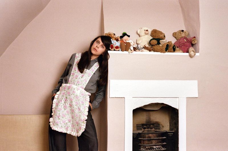 Bel Powley wearing a grey long-sleeve top and floral quilted apron, leaning against a fireplace mantel with plush teddy bears above.