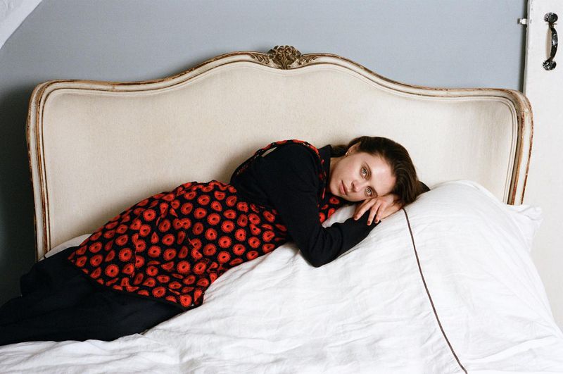 Bel Powley lying on a white bed with a curved vintage headboard wearing a red and black patterned tunic.