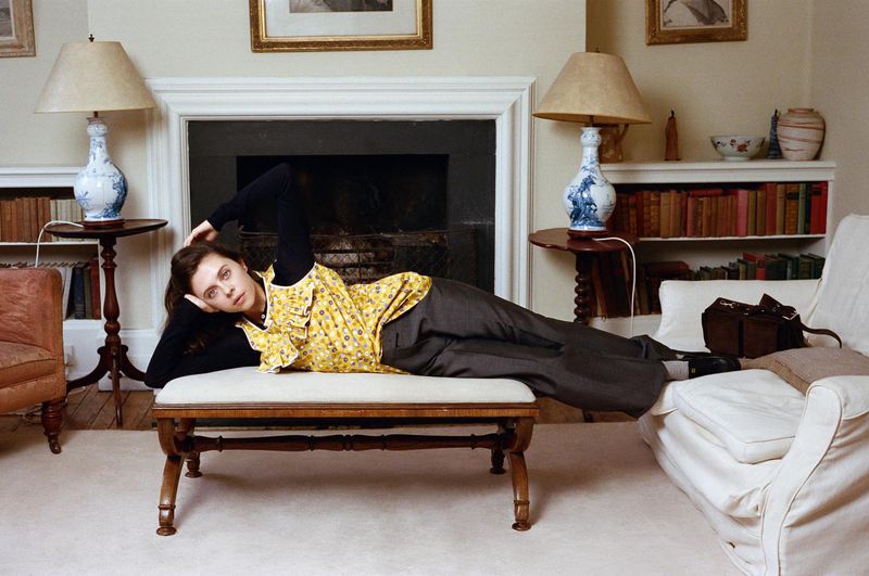 Bel Powley reclining on an upholstered bench wearing a yellow floral top and grey trousers in a living room interior.