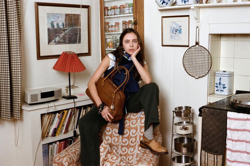 Bel Powley seated on a retro patterned armchair holding a brown leather handbag in a kitchen interior.