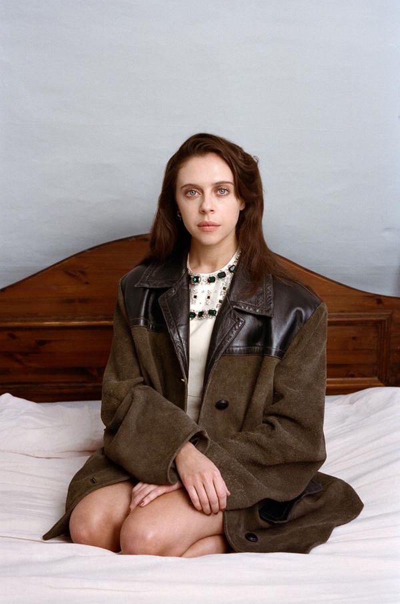 Portrait of Bel Powley kneeling on a bed wearing a brown wool coat with black leather panels for Pop Magazine.
