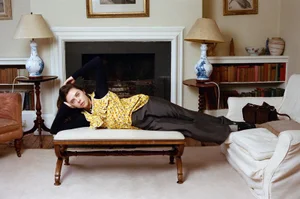 Bel Powley reclining on an upholstered bench wearing a yellow floral top and grey trousers in a living room interior.