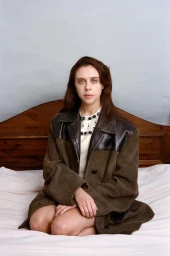 Portrait of Bel Powley kneeling on a bed wearing a brown wool coat with black leather panels for Pop Magazine.