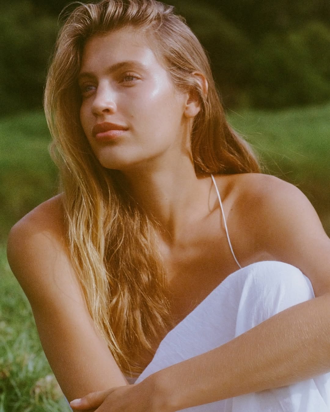 Young woman with long blonde hair, wearing a white dress, poses outdoors in a natural setting with soft, diffused lighting.