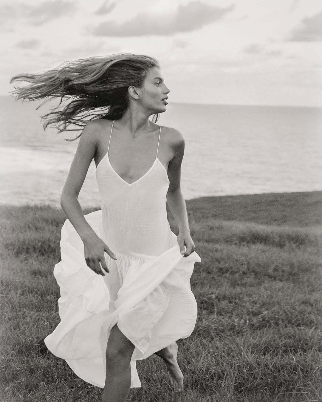 Woman in white slip dress running across a grassy coastal headland with her long hair blowing in the wind by the ocean