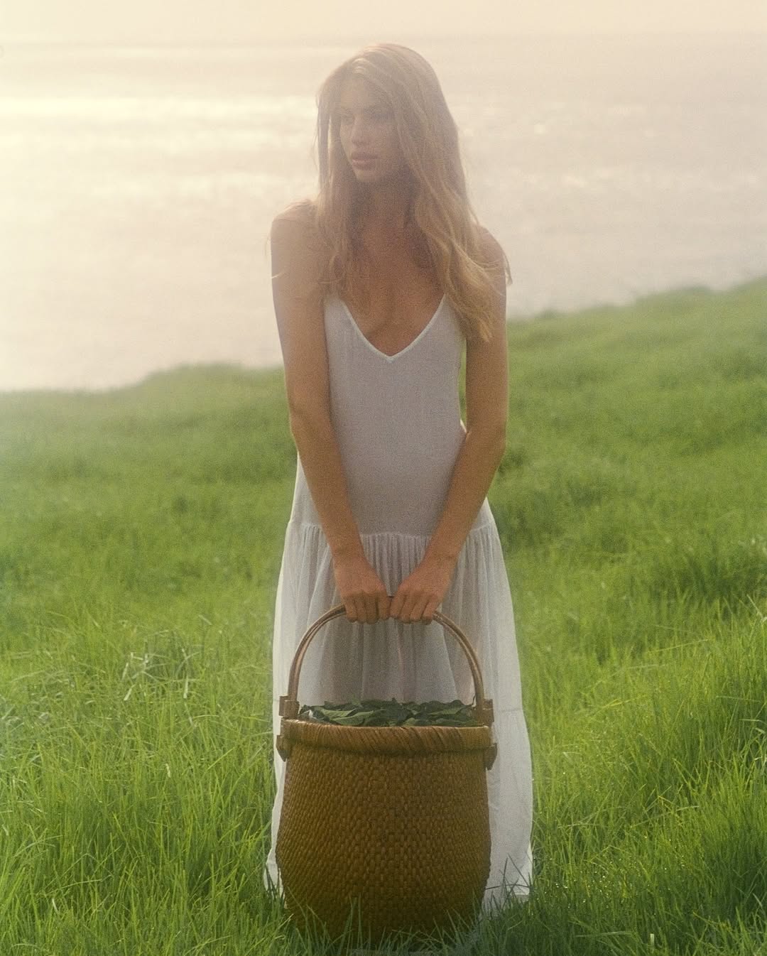 Young woman in a white dress holds a woven basket in a green field near the ocean, with soft, natural lighting enhancing the scene.