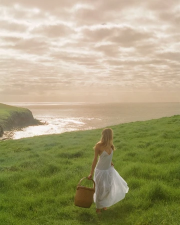 Blonde woman in a white dress carries a wicker basket walking on a grassy hill near a coastline under a cloudy sky.