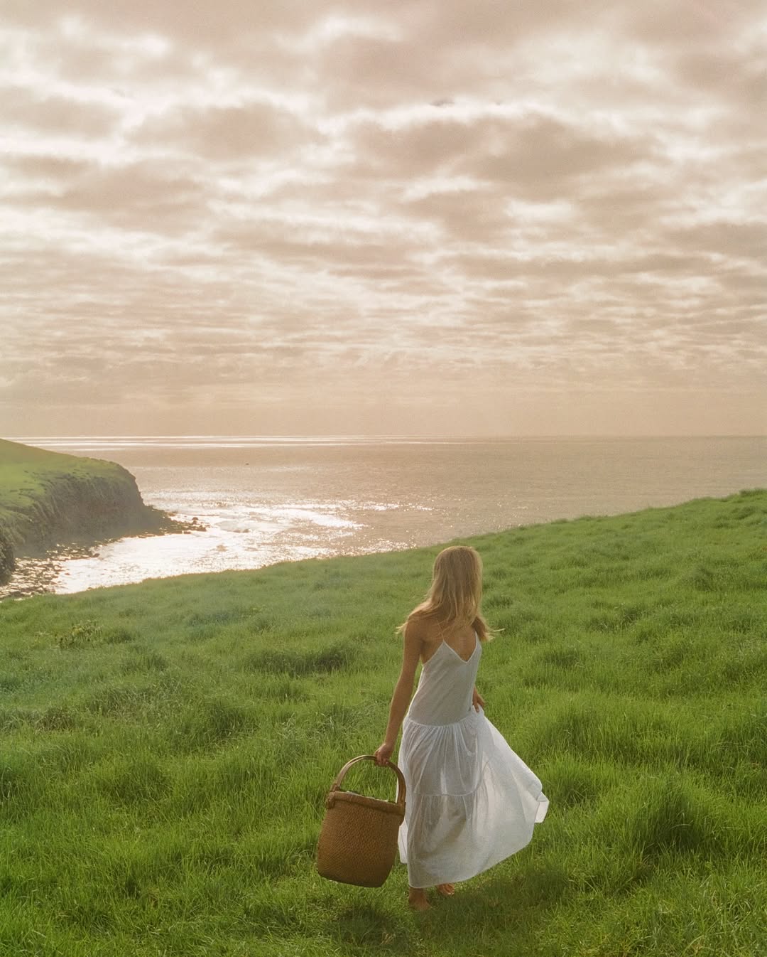 Woman in white dress walks across grassy hill with basket toward ocean under cloudy sky, creating a serene landscape.