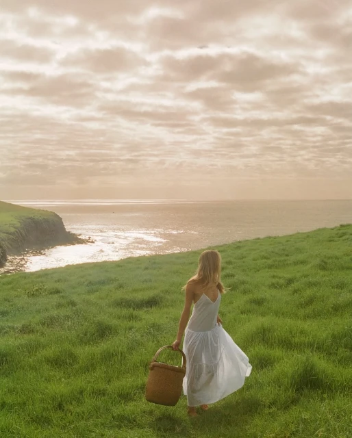 Woman in white dress walks across grassy hill with basket toward ocean under cloudy sky, creating a serene landscape.