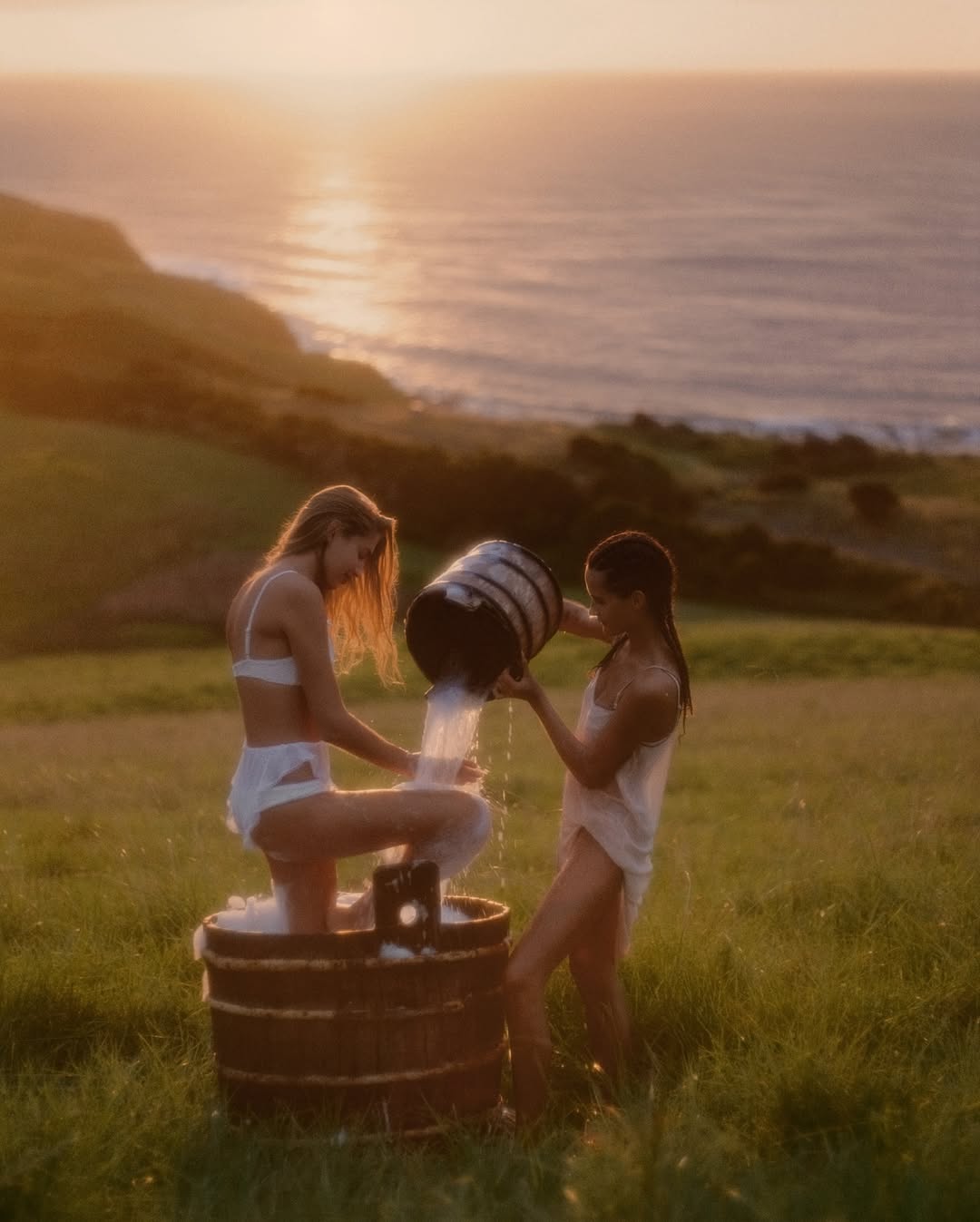 Two women in swimwear share a wooden tub in a grassy field. One pours water from a bucket, set against a coastal landscape at sunset.