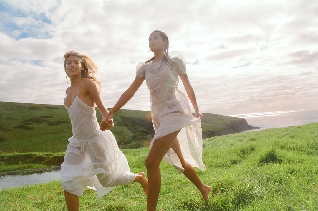 Two adult females in white dresses run hand-in-hand across a grassy field with a coastal view and cloudy sky in a sunlit outdoor scene.