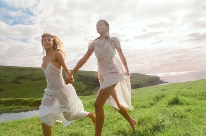 Two adult females in white dresses run hand-in-hand across a grassy field with a coastal view and cloudy sky in a sunlit outdoor scene.