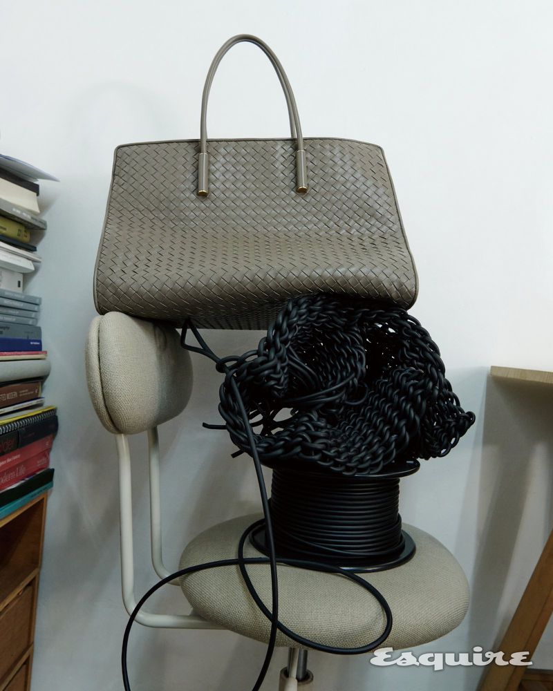 Grey woven leather tote bag resting on the backrest of a beige office chair beside black coiled cables and a stack of books.