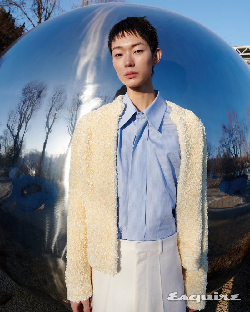 Model Minki Kwon wearing a textured cream jacket, light blue shirt, and white trousers before a large reflective sphere.