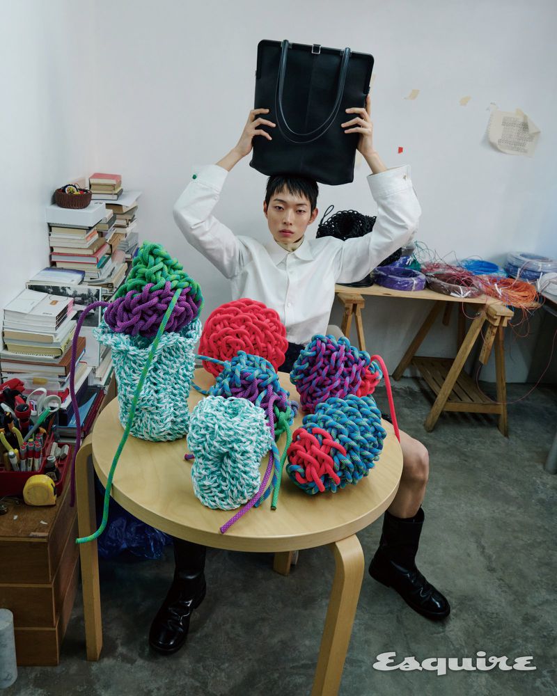 Model Minki Kwon holding a black tote bag over his head behind a table of colorful woven objects for Esquire Korea.