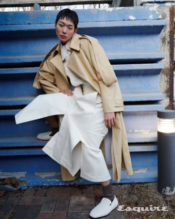 Model Minki Kwon wearing a beige trench coat and wide white trousers posed on blue outdoor steps for Esquire Korea.