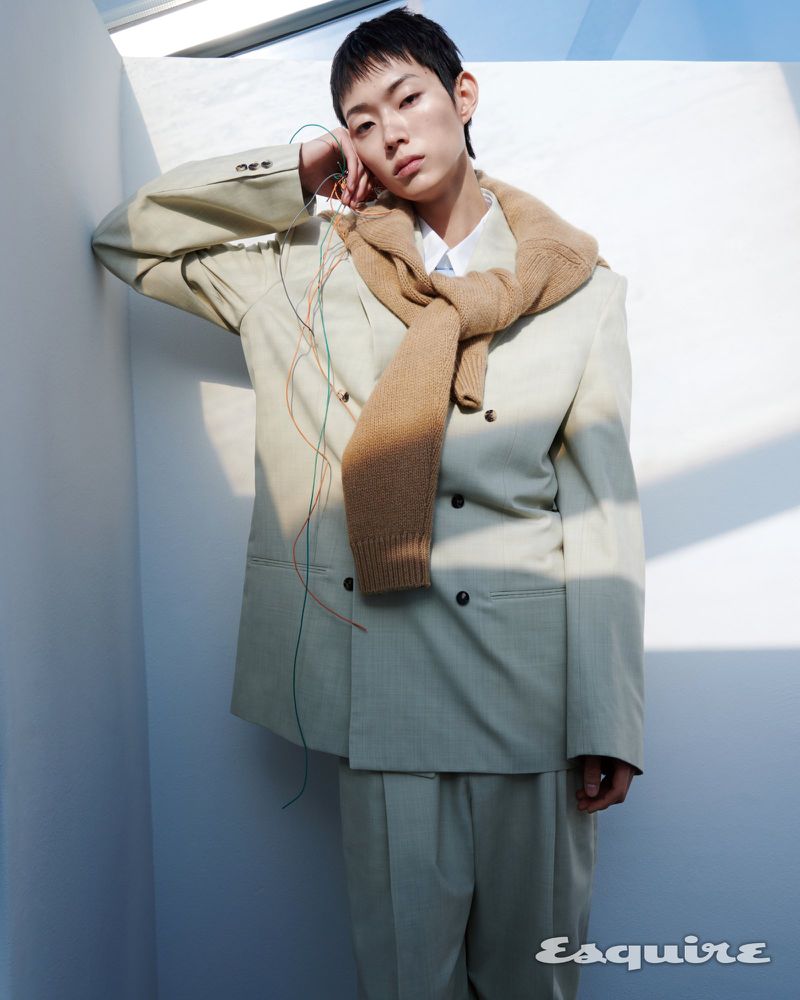 Model Minki Kwon poses in a beige double-breasted suit and camel sweater against a sunlit white corner wall for Esquire Korea.
