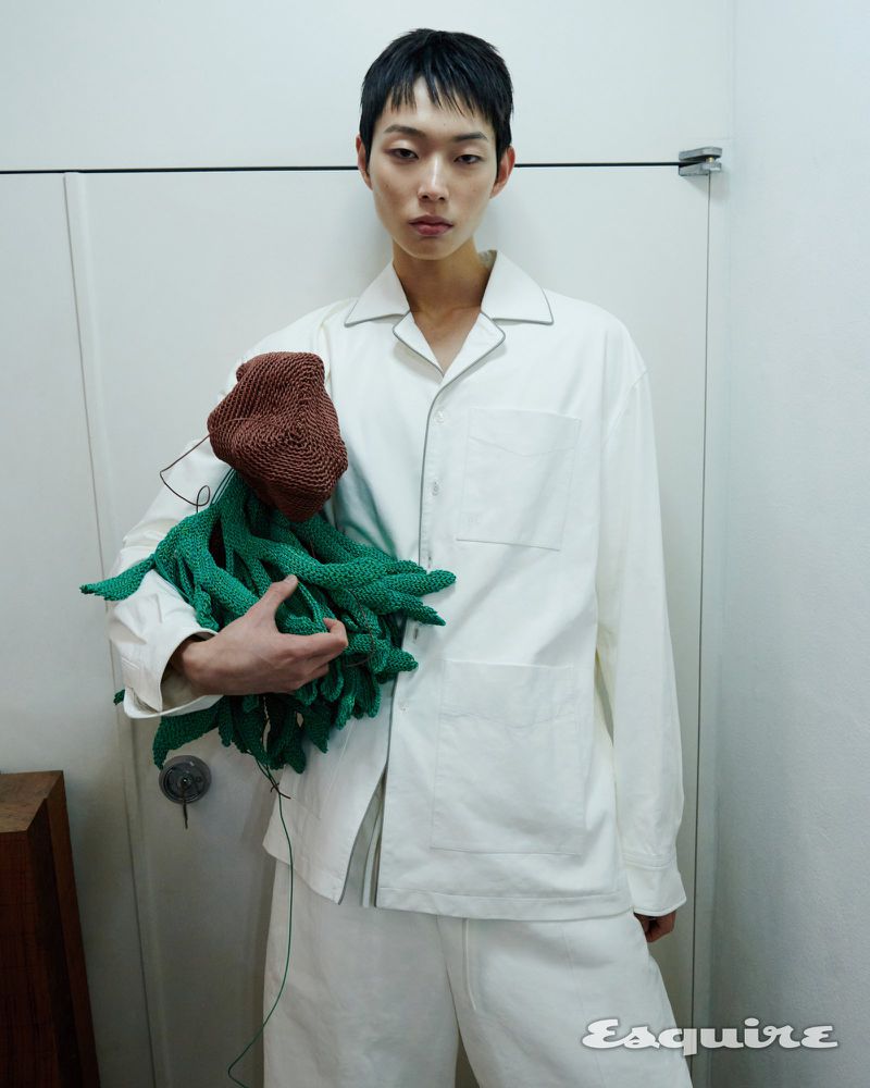 Portrait of a slender adult man with dark hair, wearing a white pajama set, holding a brown and green knit sculpture indoors.