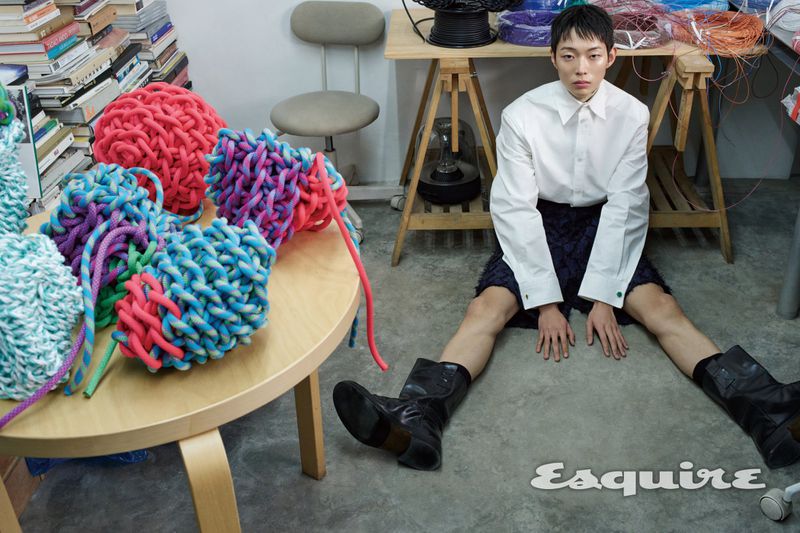 Model Minki Kwon sits on a studio floor in a white collared shirt and black boots beside woven rope art for Esquire Korea.