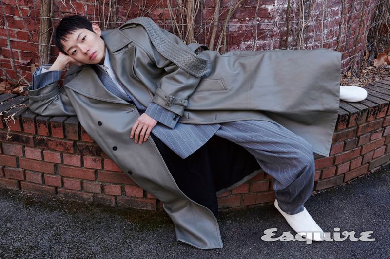 Model Minki Kwon in a long grey leather trench coat reclining on a low brick wall for an Esquire Korea editorial.
