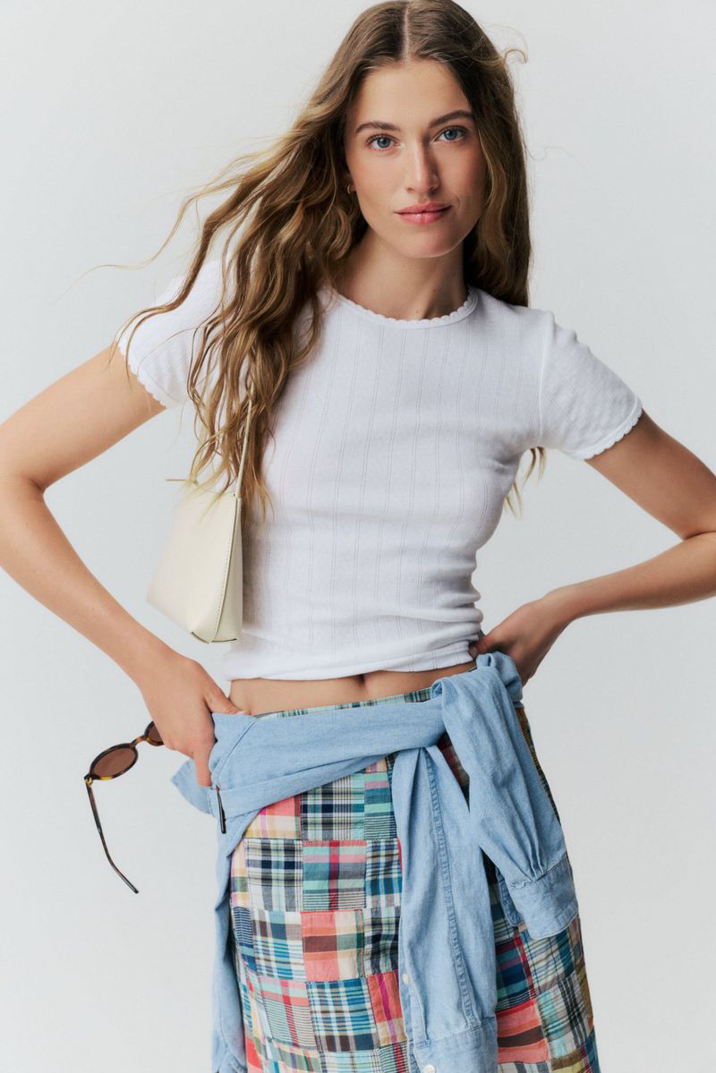 Model posing in a white ribbed t-shirt, patchwork skirt, and blue shirt tied at the waist against a neutral backdrop.