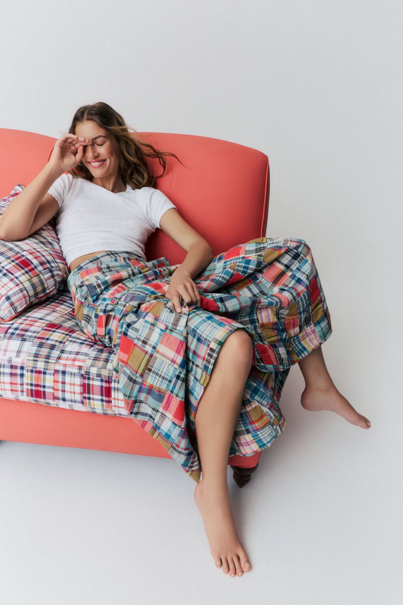 Model laughing while seated on a coral sofa wearing a white cropped t-shirt and a colorful patchwork madras skirt.