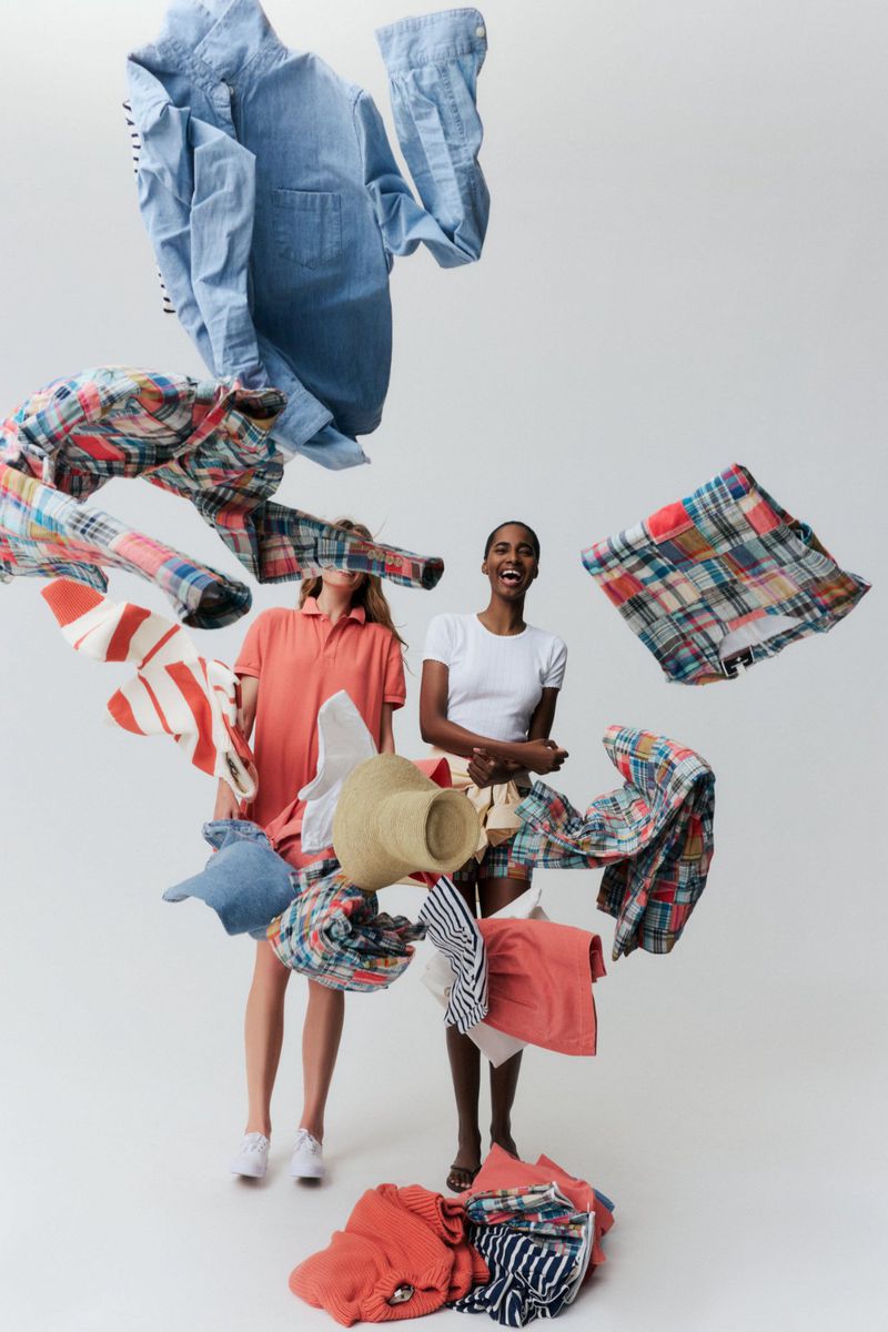 Two fashion models pose against a white seamless background surrounded by floating summer shirts and a straw hat in mid-air.